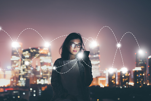 A young woman speaking on a cellphone with a city skyline in the background with abstract glowing dots and connecting dotted lines superimposed over the image